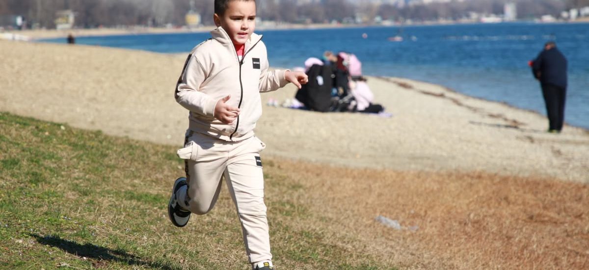 Young boy in a tracksuit runs energetically by the waterfront, embodying the joy of a sunny day outdoors.